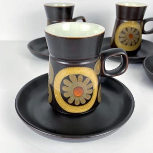 May include: A set of six vintage coffee cups and saucers. The cups are dark brown with a yellow and orange floral design. The saucers are solid black. The interior of the cups is a light green colour.