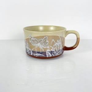 May include: A ceramic soup mug with a beige interior and handle, featuring a brown and blue exterior with a white heron and floral design. The handle is brown and beige. The mug is suitable for hot beverages.