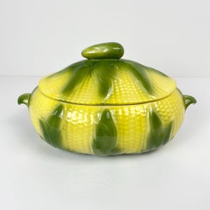 May include: A ceramic serving dish with a lid, designed to look like an ear of corn. The dish is yellow with green detailing, and has a green knob on the lid. It features two small handles on either side.