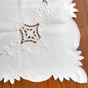 May include: A white, square embroidered linen doily with a scalloped edge. The doily features a central openwork design and floral embroidery. The intricate details and delicate stitching create a vintage aesthetic. The doily is placed on a wooden surface.
