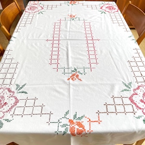 May include: White tablecloth with cross-stitch embroidery. The embroidery features a repeating geometric pattern and floral motifs in red, green, and orange. The tablecloth is rectangular and covers a wooden table.
