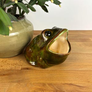 Peut inclure: Une figurine de grenouille en céramique avec un vernis brillant aux tons verts et bruns tachetés. La grenouille est représentée la bouche grande ouverte, révélant un intérieur lisse de couleur crème. La surface est réfléchissante.