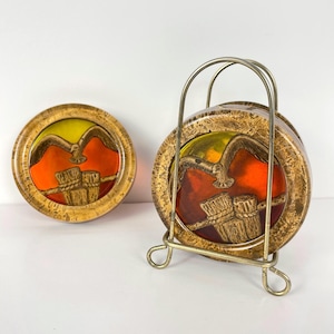 May include: Vintage coaster set featuring a bird and drum design. The coasters have a gold-coloured rim and a colourful centre with a bird and drum illustration. The set includes a metal holder for storage.