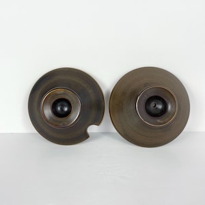 May include: Two brown ceramic lids with a dark centre and a small handle. One lid has a notch on the side. The lids have a circular shape and a matte finish. The lids are likely for a pot or other kitchenware.