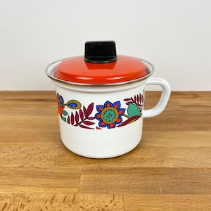 Peut inclure: Une casserole en émail blanc avec un couvercle assorti doté d'un dessus orange vif et d'une poignée noire. La casserole est décorée d'un motif floral coloré dans des tons de bleu, rouge, vert et violet, avec une anse latérale et un bord argenté.