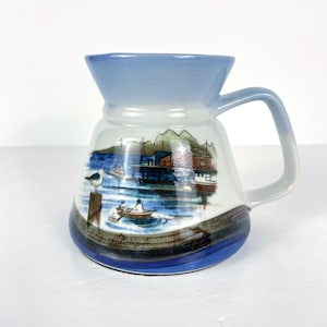 Peut inclure: Une tasse en céramique avec un bord et une anse bleu clair, représentant une scène côtière. Le motif comprend une mouette, des bateaux et des bâtiments, avec des accents bleus, bruns et blancs. La tasse a une forme évasée unique.