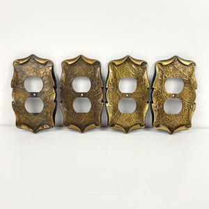 May include: Four rectangular electrical socket wall plates featuring a textured, metallic gold finish with dark brown recessed detailing. Each plate has a decorative, scalloped edge and two circular openings for a standard duplex wall socket.