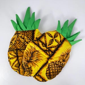 Peut inclure: Deux dessous de plat en forme d'ananas. Les dessous de plat ont un fond jaune avec un motif géométrique marron et doré. Le dessus des dessous de plat est vert, ressemblant à des feuilles d'ananas. Les dessous de plat sont sur fond blanc.