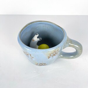 May include: A light blue ceramic mug with a whimsical snail figurine inside. The mug has a floral design and a curved handle. The snail has a white body and yellow shell. A unique and decorative item.