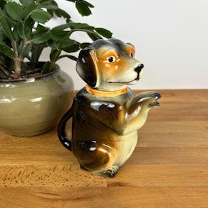 May include: A ceramic creamer shaped like a seated dog with brown, black, and tan colouring. The dog has a glossy finish, a yellow collar, and a handle formed by its tail. One paw is extended forward, and the head is turned slightly to the side.