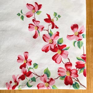 May include: White fabric with a floral pattern featuring pink and red flowers with yellow centres and green leaves. The flowers are arranged on brown stems, creating a decorative design.