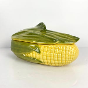 May include: A ceramic corn-shaped container with a lid. The container is yellow with a textured surface, mimicking corn kernels. The lid is green, resembling corn husks. The design is a decorative kitchen item.