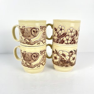 May include: Four beige ceramic mugs stacked together. Two mugs feature a brown owl design perched on a branch, while the other two display a floral pattern in brown. Each mug has a curved handle.