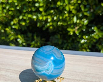May include: A polished blue agate sphere with white and light blue swirls, presented on a decorative gold-coloured metal stand. The sphere is a decorative item, suitable for home decor or display.