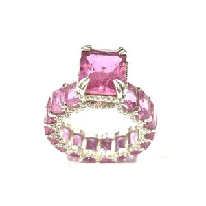 Può includere: Un anello d'argento con una grande gemma rosa rettangolare al centro, circondata da gemme rosa più piccole. La fascia dell'anello è anch'essa ornata di gemme rosa, creando un design continuo e scintillante. L'anello è su sfondo bianco.