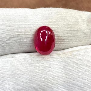Puede incluir: Una gema de rubí pulida, de forma ovalada. La gema es de un color rojo intenso y saturado, con una superficie lisa y reflectante. El rubí se encuentra sobre un fondo de tela blanca, resaltando su vibrante tonalidad y claridad.