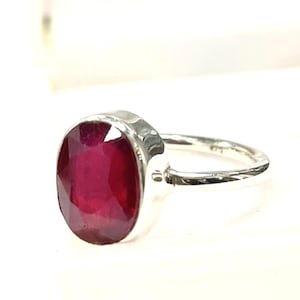 May include: A silver ring featuring a large, oval-cut ruby gemstone. The ruby is a deep, vibrant red color, set in a textured silver band. The ring's design is simple yet elegant, showcasing the gemstone's natural beauty.