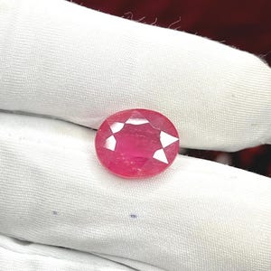 May include: A faceted, oval-shaped ruby gemstone. The gem is a vibrant red colour with sharp, white facets that catch the light. The ruby is set against a white fabric background, highlighting its colour and clarity.