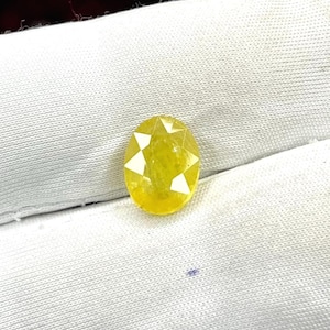 Op de afbeelding: Ovaalvormige gele edelsteen met witte stervormige facetten. De edelsteen ligt tegen een witte stoffen achtergrond. De slijping creëert een steruitbarsting-effect, waardoor de glans wordt versterkt. Geschikt voor sieraden.