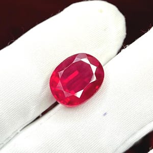 Può includere: Una gemma di rubino sfaccettata di forma ovale. La gemma è di un colore rosso vibrante ed è posta su uno sfondo bianco. Le sfaccettature creano un effetto scintillante, evidenziando la chiarezza e la brillantezza della pietra. Il rubino è una gemma classica.