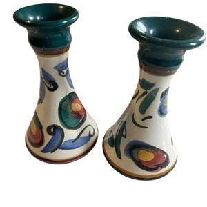 May include: A pair of ceramic candle holders with a white base and colourful abstract designs. The holders have a green top and a brown rim.