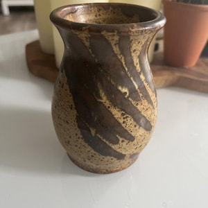 May include: A small, handcrafted ceramic vase with a speckled beige base and dark brown diagonal stripes. The vase has a slightly flared rim and a rounded body, ideal for displaying small flowers or decorative branches.