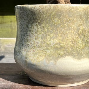 Może przedstawiać: Ceramiczna donica z unikalnym, teksturowanym szkliwem. Donica ma zaokrąglony kształt z szerokim otworem. Szkliwo ma nakrapiany wygląd z odcieniami zieleni, beżu i bieli, tworząc naturalną, ziemistą estetykę.