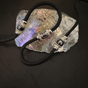 May include: A black cord necklace featuring three silver-toned beads. Each bead is cylindrical with a central dark stone and small clear crystals on the ends. The center bead displays a small heart detail. The cord is smooth and woven in texture.