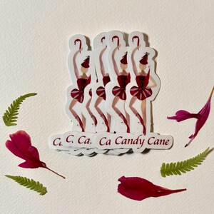 May include: A stack of five white stickers with a red and white illustration of a woman in a red dress holding a candy cane. The text "Ca Ca Ca Ca Candy Cane" is printed on the bottom of the stickers.