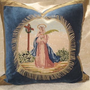 以下が含まれることがあります： 金色のトリムが付いた青いベルベットの枕には、十字架の前に立っているヤシの葉を持った女性が針仕事で描かれています。