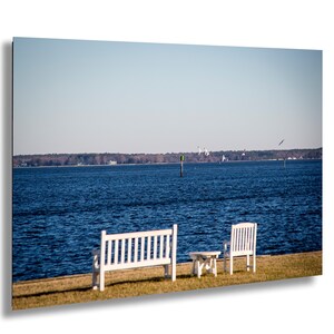 May include: A serene outdoor scene featuring a white bench, chair, and small table set on a grassy area. The backdrop is a vast expanse of blue water under a clear sky, with a distant treeline and several birds in flight.