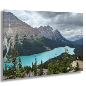 Peut inclure: Photographie de paysage d'un lac turquoise entouré de montagnes et de forêts de conifères. Le ciel est couvert de nuages gris. Quelques personnes sont visibles sur une plateforme d'observation.