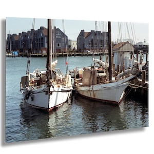 Può includere: Due barche a vela bianche ormeggiate fianco a fianco in un porto. Le barche hanno alberi e sartiame in legno, con una striscia rossa lungo lo scafo. Edifici con tetti grigi sullo sfondo e l'acqua è blu scuro.