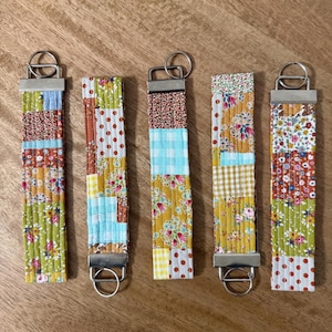 May include: Five colorful fabric key fobs with silver-toned hardware. Each fob features a patchwork design with floral, polka dot, and gingham patterns in various colors, including green, yellow, blue, and brown. The key fobs are arranged on a wooden surface.