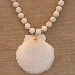 May include: A necklace featuring a series of matte white spherical beads separated by small gold-toned spacers. The centerpiece is a large, natural white scallop shell pendant adorned with a single small, round, clear crystal set in gold.