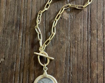 May include: Gold-toned necklace with a large link chain and a toggle clasp. The pendant features a silver-colored coin with an elephant design, set within a gold-colored, ornate frame. The necklace rests on a wooden surface.