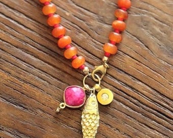 Può includere: Una collana con una fila di perline arancioni rotonde infilate su un cordino rosso. La collana include una chiusura a moschettone color oro con tre ciondoli pendenti: una pietra rosa quadrata, un pesce color oro testurizzato e un piccolo disco dorato.