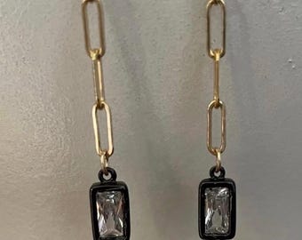 May include: A pair of gold-toned chain earrings. Each earring features a rectangular, clear gemstone set in a black frame, suspended from a gold chain. A small gold ball hangs below the gemstone.