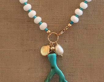 Può includere: Una collana con perline bianche e turchesi, con chiusura e ciondolo dorati. Il ciondolo include un ramo di corallo turchese, una conchiglia dorata e una perla bianca. La collana è esposta su uno sfondo neutro.