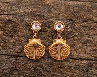 Può includere: Un paio di orecchini a forma di conchiglia color oro con una gemma rotonda e trasparente sulla parte superiore di ogni orecchino. Gli orecchini pendono da una piccola catena d'oro. Sfondo in legno.