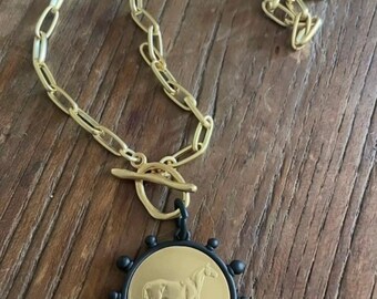 May include: Gold-tone necklace with a horse pendant. The pendant features a gold-colored horse design on a black circular frame with small black spheres around the edge. The necklace has a gold chain and a toggle clasp.