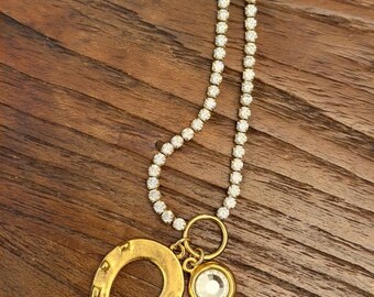 Può includere: Una collana dorata con un ciondolo a forma di ferro di cavallo e un pendente rotondo di cristallo trasparente. La collana ha una delicata catena di piccoli cristalli scintillanti. Il ciondolo a forma di ferro di cavallo ha una superficie strutturata.