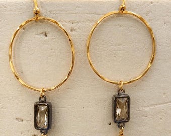 May include: A pair of gold-tone hoop earrings with a hammered texture. Each earring features a rectangular, smoky-colored gemstone pendant set in a dark metal frame, and a small gold ball at the bottom.
