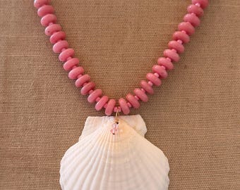 Può includere: Una collana con un grande ciondolo a forma di conchiglia bianca e una catena di perline rosa. La collana ha una chiusura color oro e un piccolo ciondolo rosa appeso alla conchiglia. Le perline sono rosa chiaro.