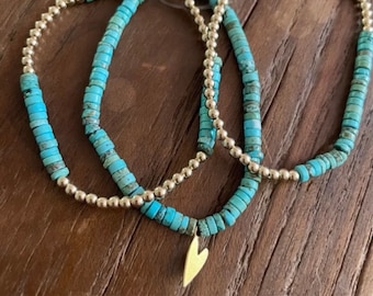 Può includere: Collana a tre fili con perline a disco turchese e piccole perline dorate. La collana ha un ciondolo a forma di cuore dorato. I gioielli sono esposti su una superficie di legno.