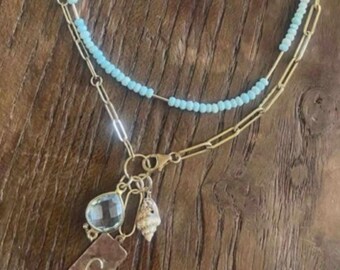 May include: A layered necklace featuring a strand of light blue beads and a gold-toned paperclip chain. The chain includes three charms: a clear teardrop-shaped stone, a small textured seashell, and a rectangular gold-toned tag engraved with the letter C.