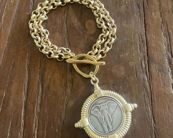 May include: Gold-toned bracelet with a toggle clasp and a pendant featuring an elephant design. The pendant is round with a silver-colored elephant image in the center, surrounded by a gold-colored frame. The bracelet has a triple-strand chain.