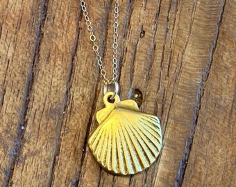 May include: A gold-colored seashell pendant necklace. The pendant is a detailed, textured seashell shape, suspended from a delicate gold chain. The necklace is displayed on a wooden surface.