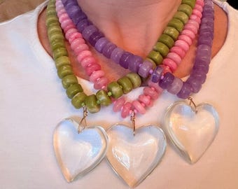 Può includere: Tre collane con ciondoli a forma di cuore. Le collane presentano perline nelle tonalità del verde, del rosa e del viola. I ciondoli a forma di cuore sono trasparenti. Le collane sono appese su uno sfondo bianco.