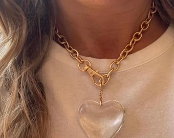Può includere: Collana a catena color oro con un grande ciondolo a forma di cuore trasparente. La collana presenta un design a catena grossa e una chiusura a moschettone. Il ciondolo a forma di cuore è trasparente e pende da un delicato filo d'oro.